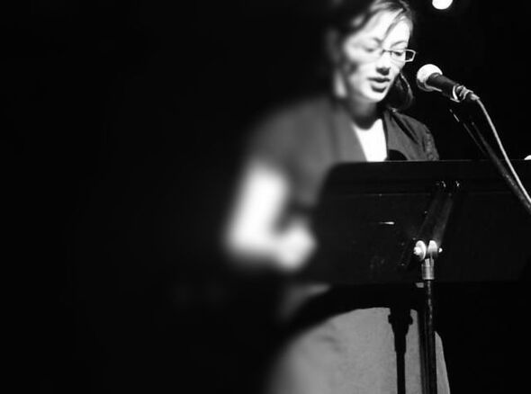 A black and white photo of Sarah, a white woman with brown hair. The hair is pulled back, with some hair in her face. She wears large, dangling triangle earrings and rectangle glasses. She wears a black dress. She stands at a microphone, looking down at a music stand. 
