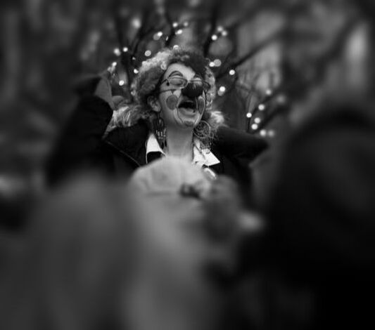 A black and white photo. In the center of it is a white woman dressed as a clown, shouting something to a gathered crowd. Other than the woman at the center, the picture is blurry.